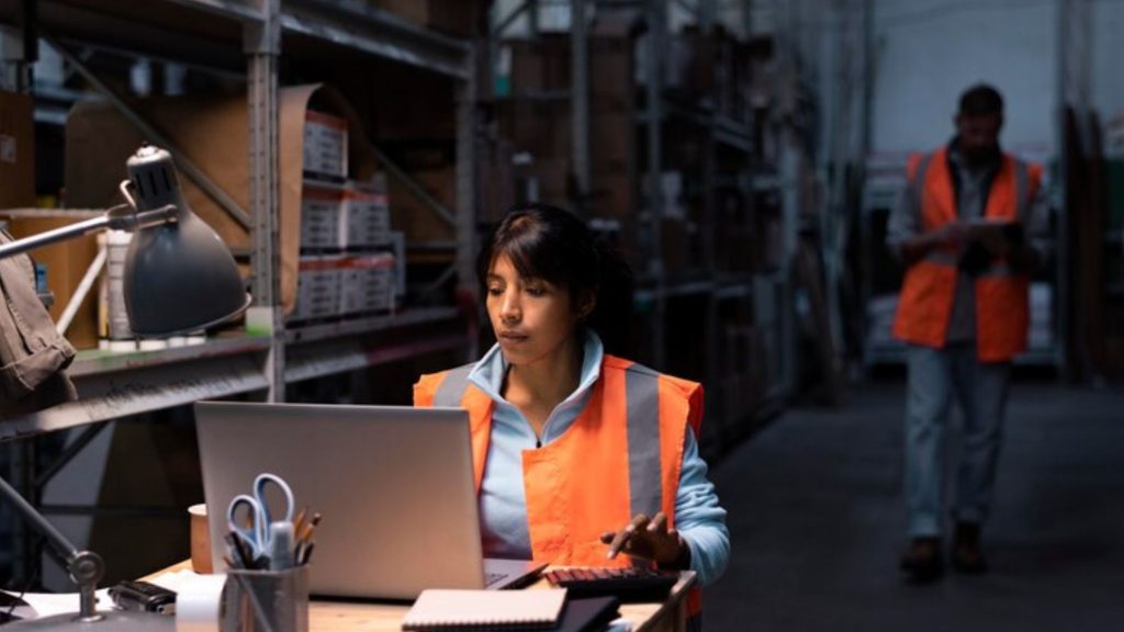 A woman in an orange vest diligently works on a laptop, focused and engaged in answering complaints of a logistic industry